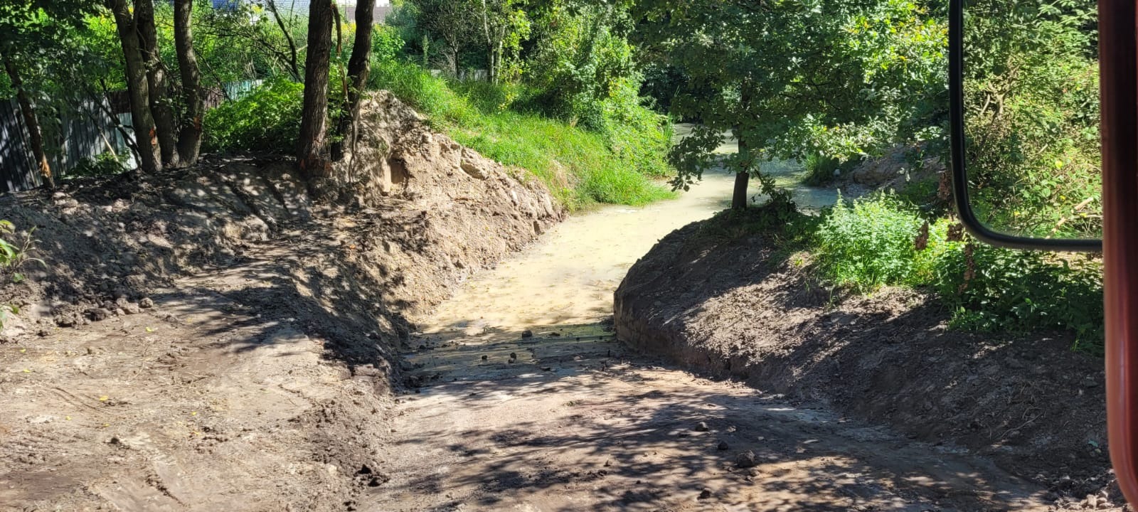 Wykopany rów melioracyjny z wodą, otoczony ziemią i zielenią. Widok z kabiny maszyny, z fragmentem lusterka po prawej stronie kadru. Słoneczny dzień.