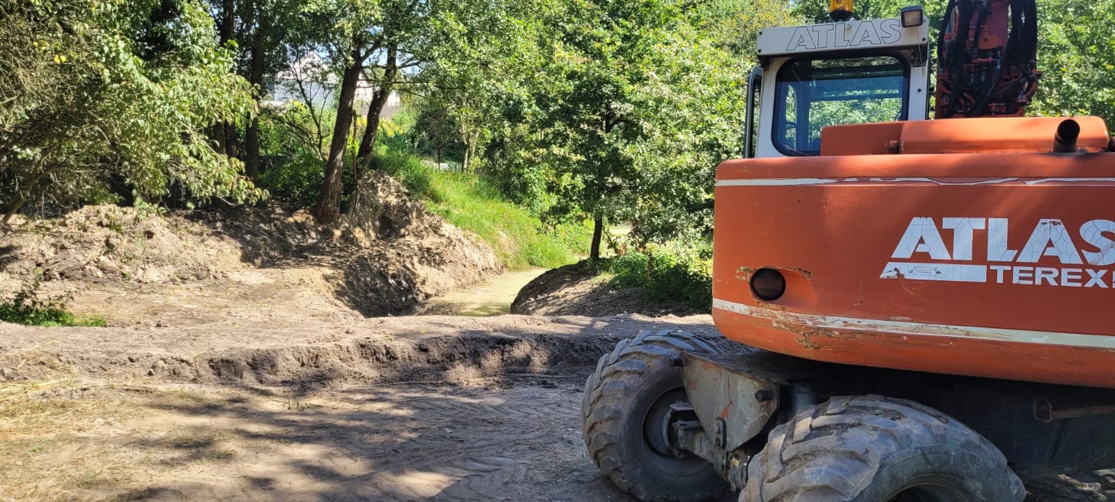 Pomarańczowa koparka Atlas na tle wykopanego rowu z wodą, otoczonego zielenią. Widoczne ślady opon i świeżo naruszona ziemia.