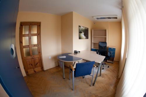 Jasne biuro z drewnianą podłogą w jodełkę, okrągłym stołem z niebieskim blatem, niebieskim krzesłem i biurkiem z czarnym krzesłem obrotowym w tle, widziane przez szerokokątny obiektyw.