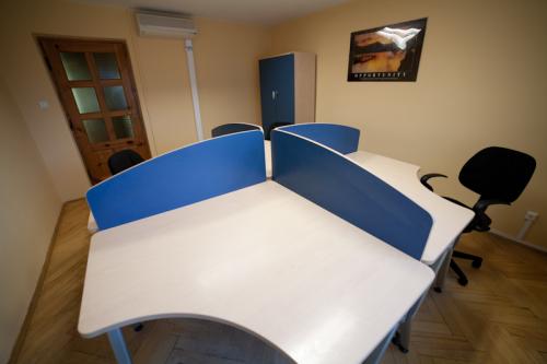 Wnętrze biura z czterema stanowiskami pracy oddzielonymi niebieskimi przegrodami, krzesło biurowe, drewniana podłoga, plakat na ścianie.