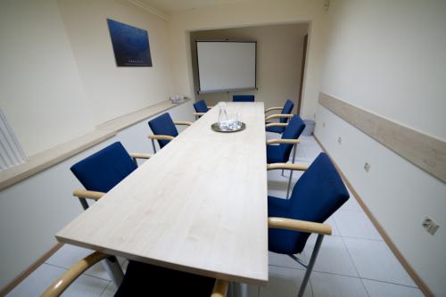 Jasna sala konferencyjna z długim stołem z drewna, niebieskimi krzesłami i ekranem projekcyjnym w tle.