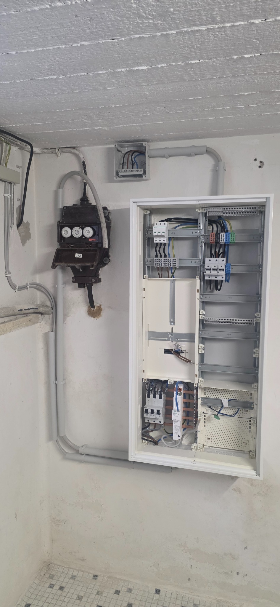 Otwarta, biała skrzynka rozdzielcza z okablowaniem na ścianie, obok stara skrzynka z bezpiecznikami topikowymi. Szare rury instalacyjne na ścianie.
