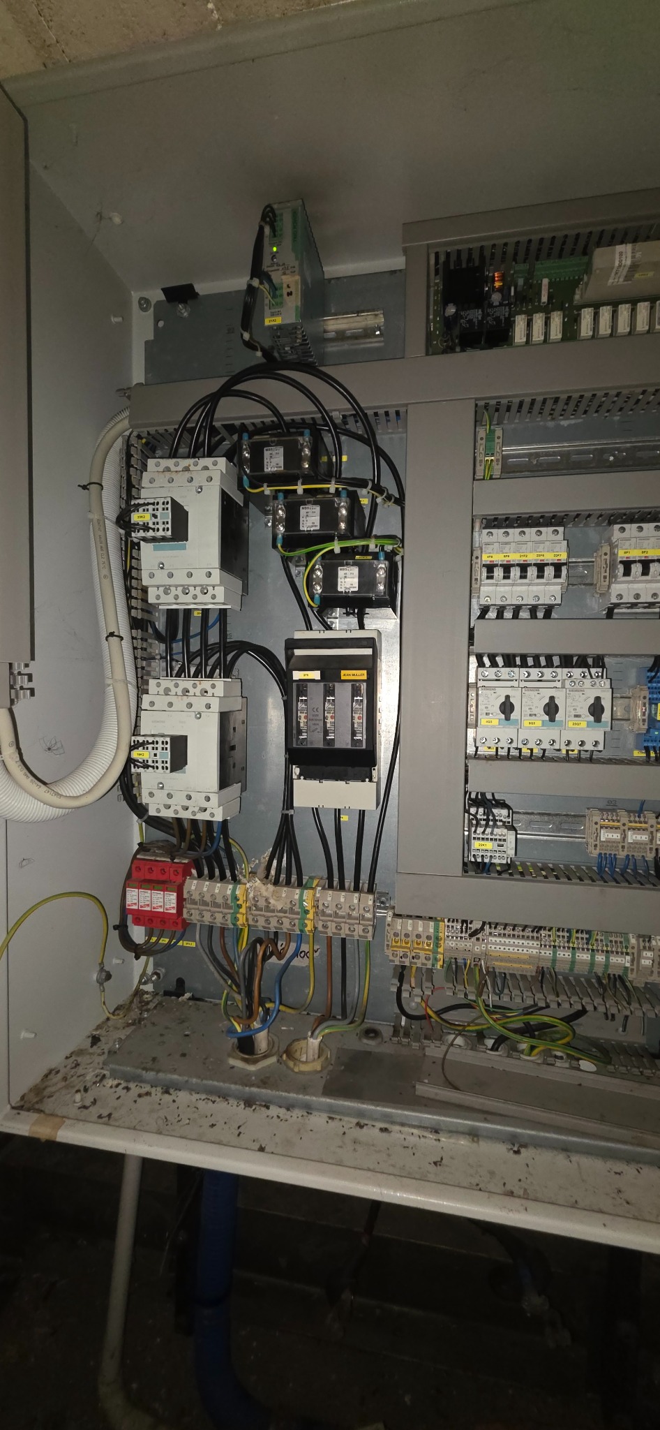 Otwarta szafa elektryczna z widocznymi stycznikami, bezpiecznikami i przewodami. Instalacja w szarej obudowie, widoczne elementy sterowania i zasilania.