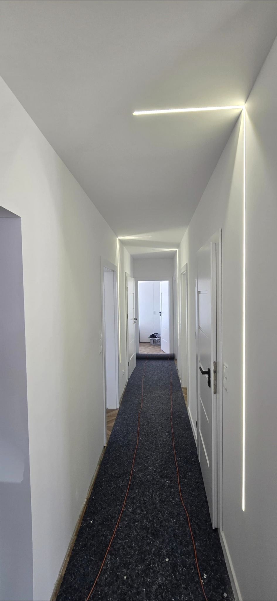 Nowoczesny korytarz z białymi ścianami i liniowym oświetleniem LED wzdłuż sufitu i ścian, tworzącym geometryczny wzór. Podłoga zabezpieczona przed zabrudzeniem.