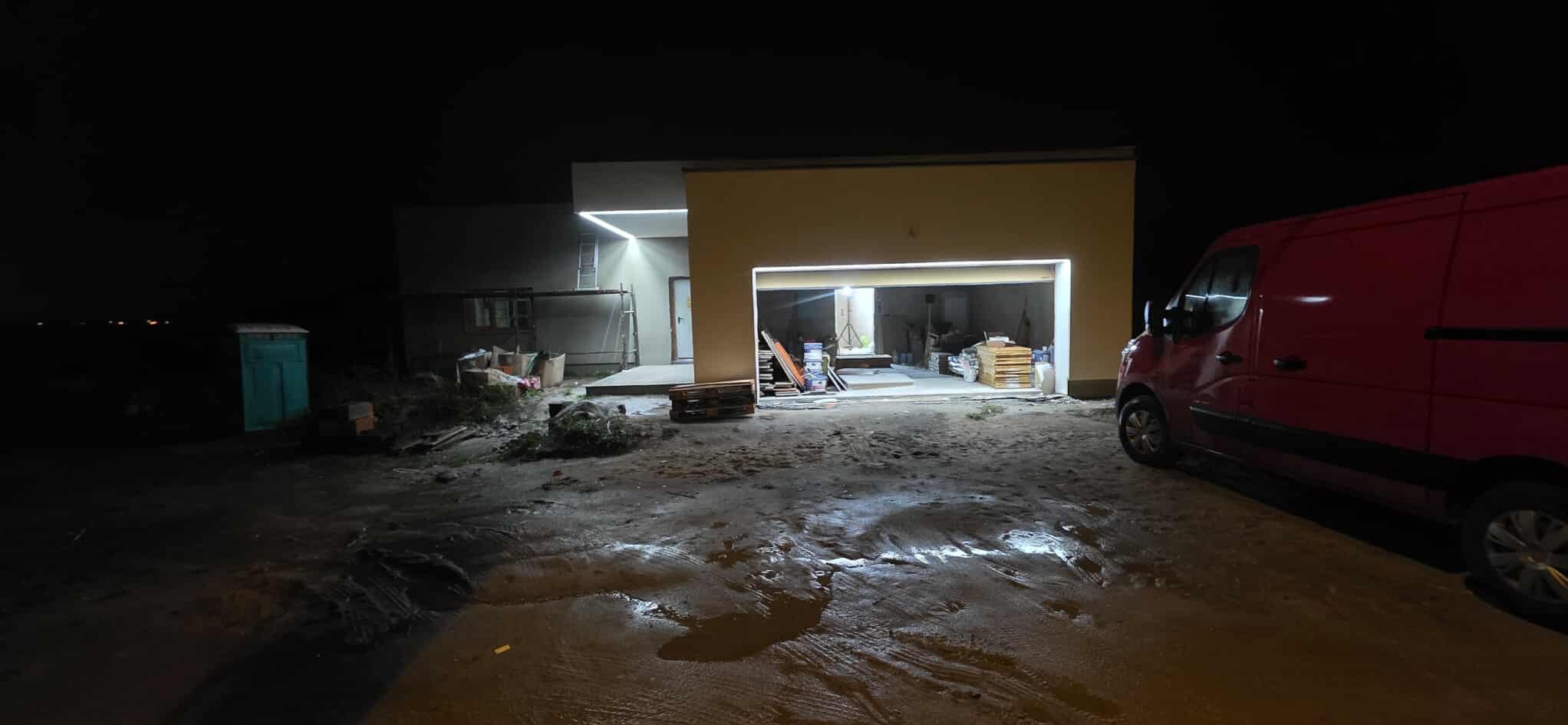 Nocne ujęcie domu z oświetleniem zewnętrznym LED, widoczny samochód dostawczy i teren budowy. Oświetlenie podkreśla nowoczesną architekturę budynku.