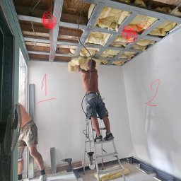 VAN GLOBAL SPÓŁKA Z OGRANICZONĄ ODPOWIEDZIALNOŚCIĄ - Montaż izolacji stropu: mężczyzna na drabinie umieszcza wełnę mineralną w metalowej konstrukcji sufitu podwieszanego. Widoczne narzędzia i fragmenty izolacji na podłodze.
