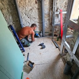 VAN GLOBAL SPÓŁKA Z OGRANICZONĄ ODPOWIEDZIALNOŚCIĄ - Hydraulik w trakcie pracy nad instalacją w pomieszczeniu z surowymi ścianami. Widoczne rury, narzędzia i konstrukcja szkieletowa ściany.