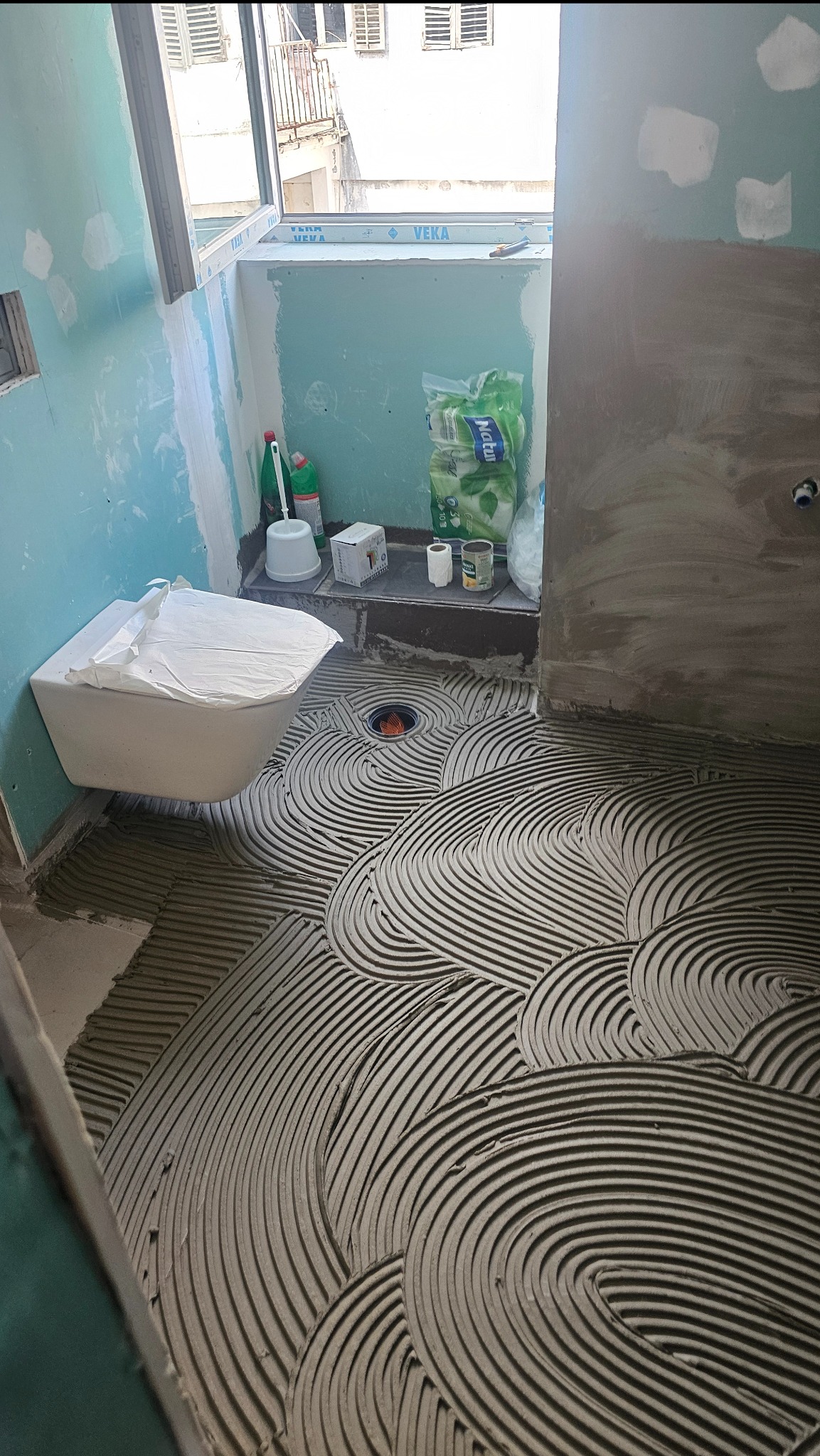 Przygotowanie podłoża pod płytki w łazience: warstwa kleju ułożona w charakterystyczne półokręgi, widoczny odpływ liniowy i fragment toalety podwieszonej.