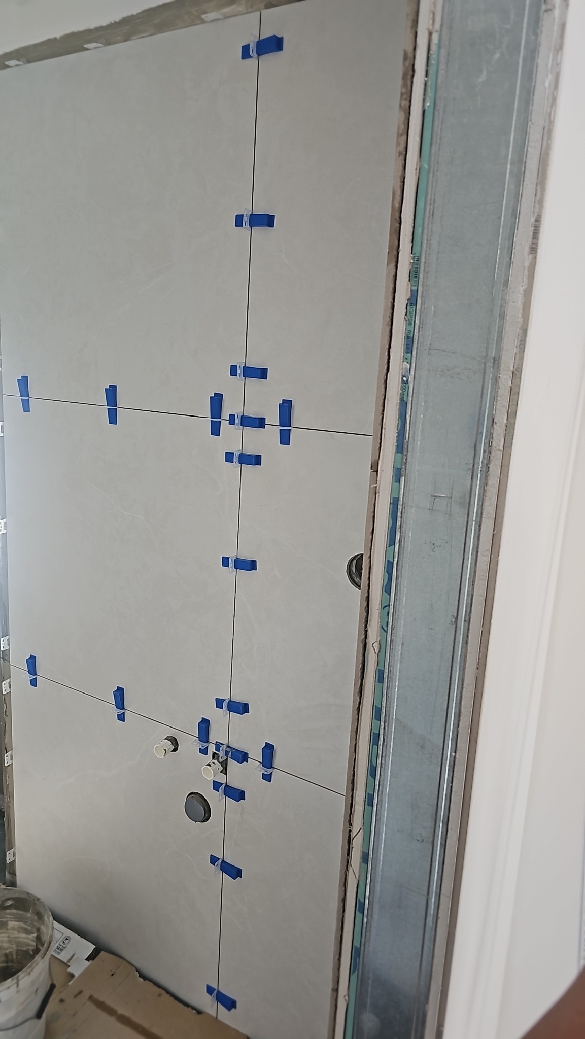 Ściana z jasnych płytek, z niebieskimi klipsami poziomującymi i czarnymi liniami fug, widoczne rury i metalowa rama, etap prac wykończeniowych w łazience.