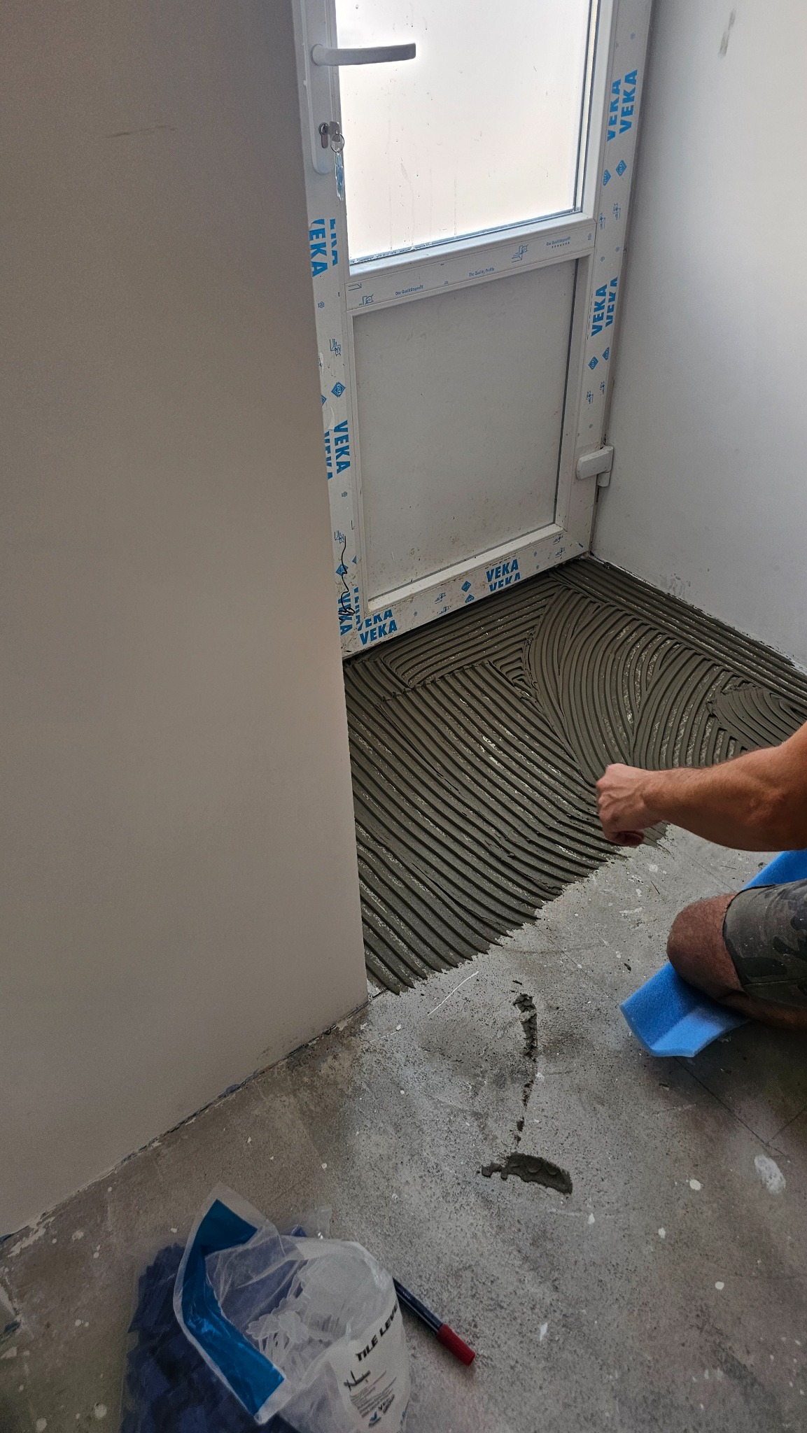 Przygotowanie podłoża pod płytki w małym pomieszczeniu z drzwiami. Widoczna warstwa kleju rozprowadzona pacą ząbkowaną. W tle torba z materiałami budowlanymi.