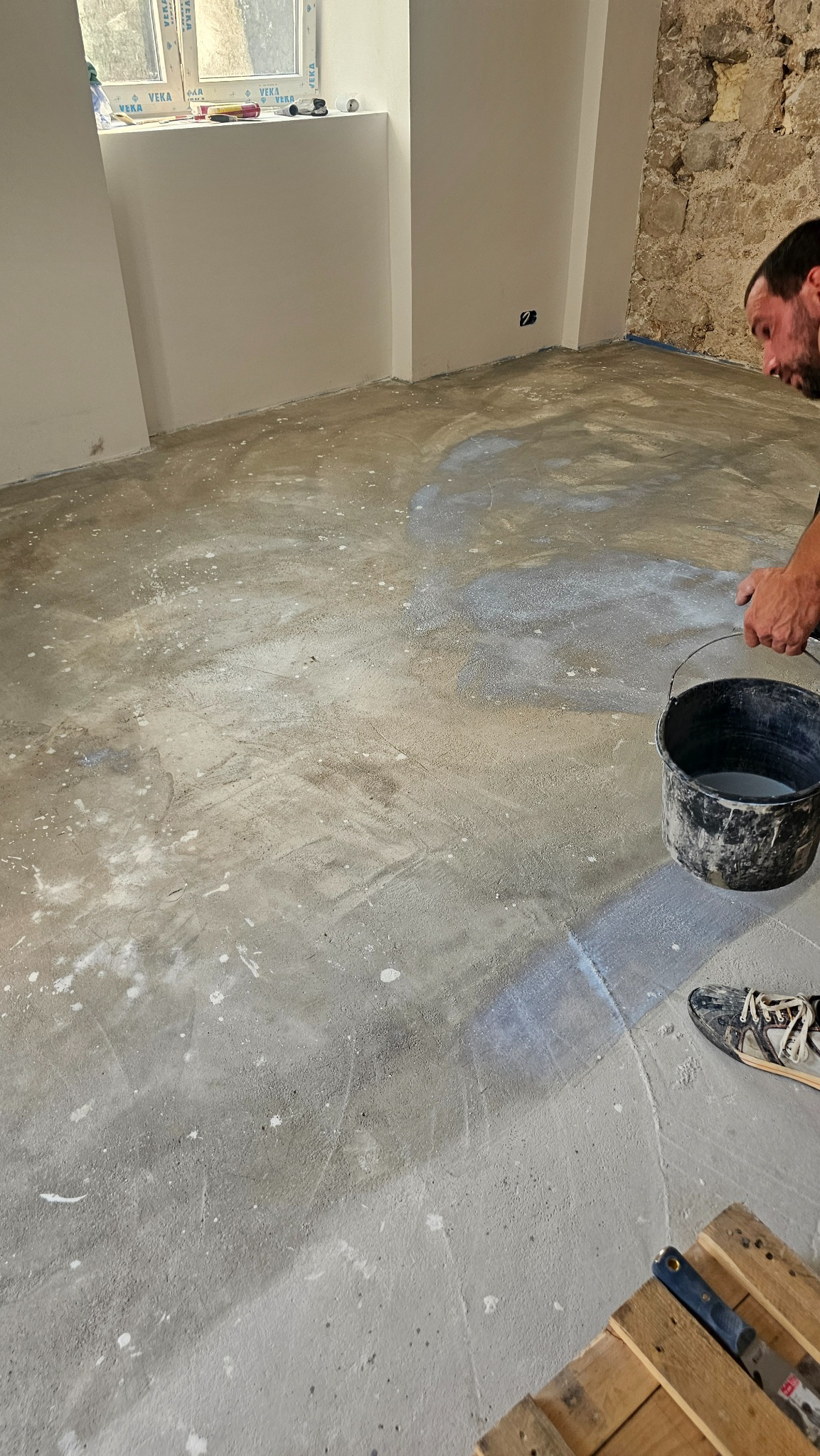 Wyrównywanie podłoża szpachlą. Widoczny fragment ściany z kamienia, okno i narzędzia. Mężczyzna z wiadrem w trakcie pracy. Podłoże pokryte szpachlą.