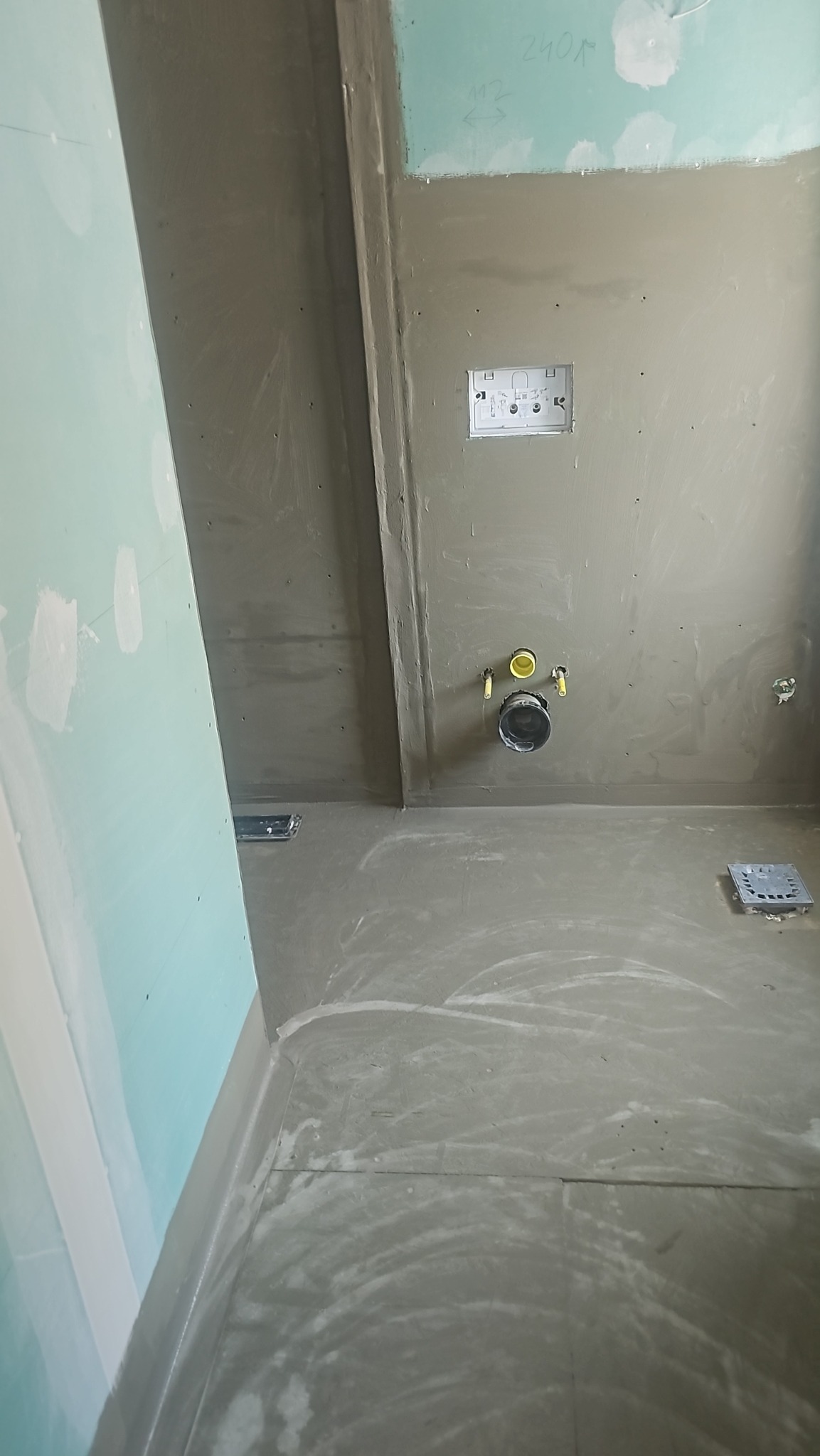 Przygotowana do wykończenia łazienka z odpływem liniowym i punktowym. Ściany pokryte szarą hydroizolacją, widoczne instalacje. Minimalistyczny design.