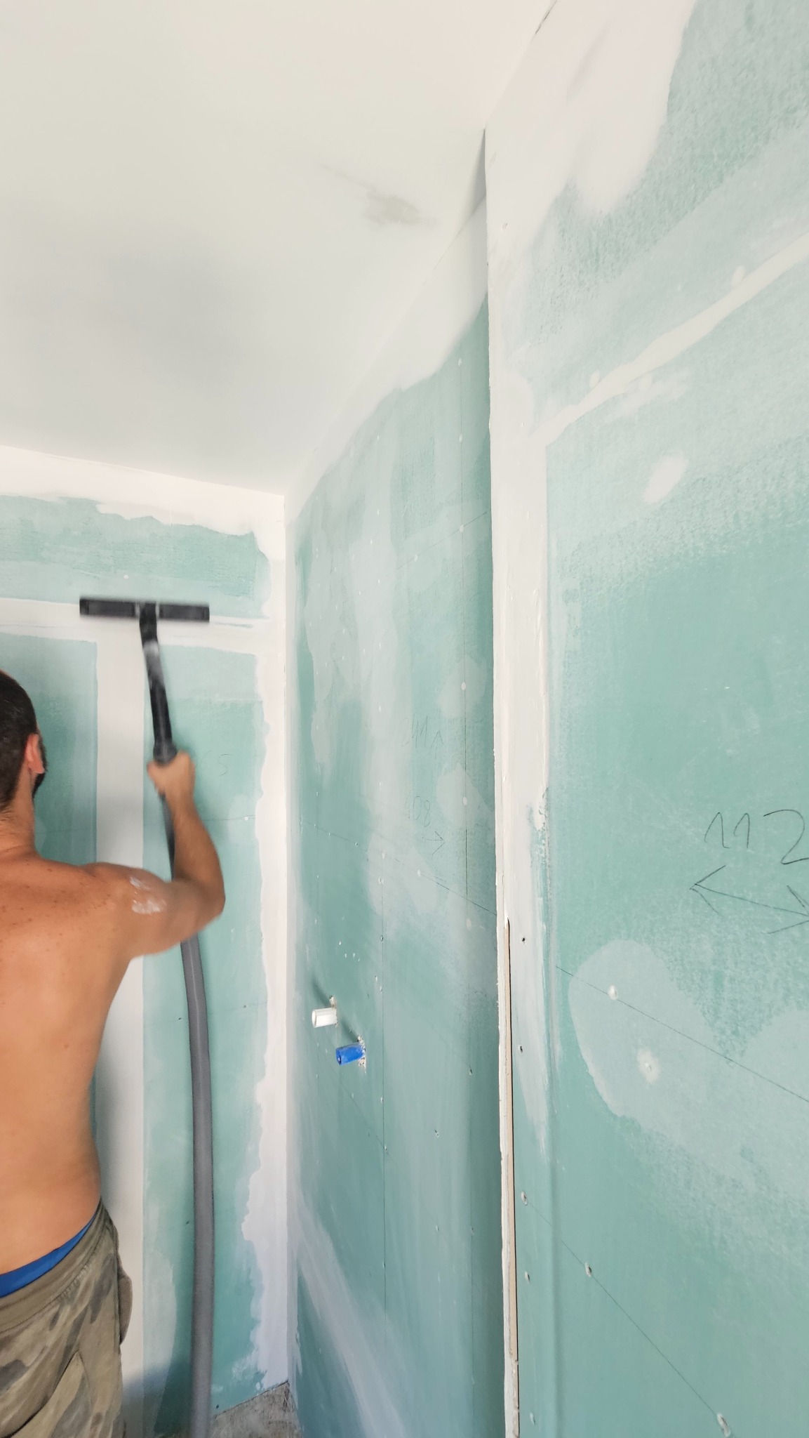 Gładzenie ściany z płyt gipsowo-kartonowych w łazience. Widoczne niebieskie i białe zaślepki instalacji wodnej. Mężczyzna w trakcie pracy, zacieranie łączeń.