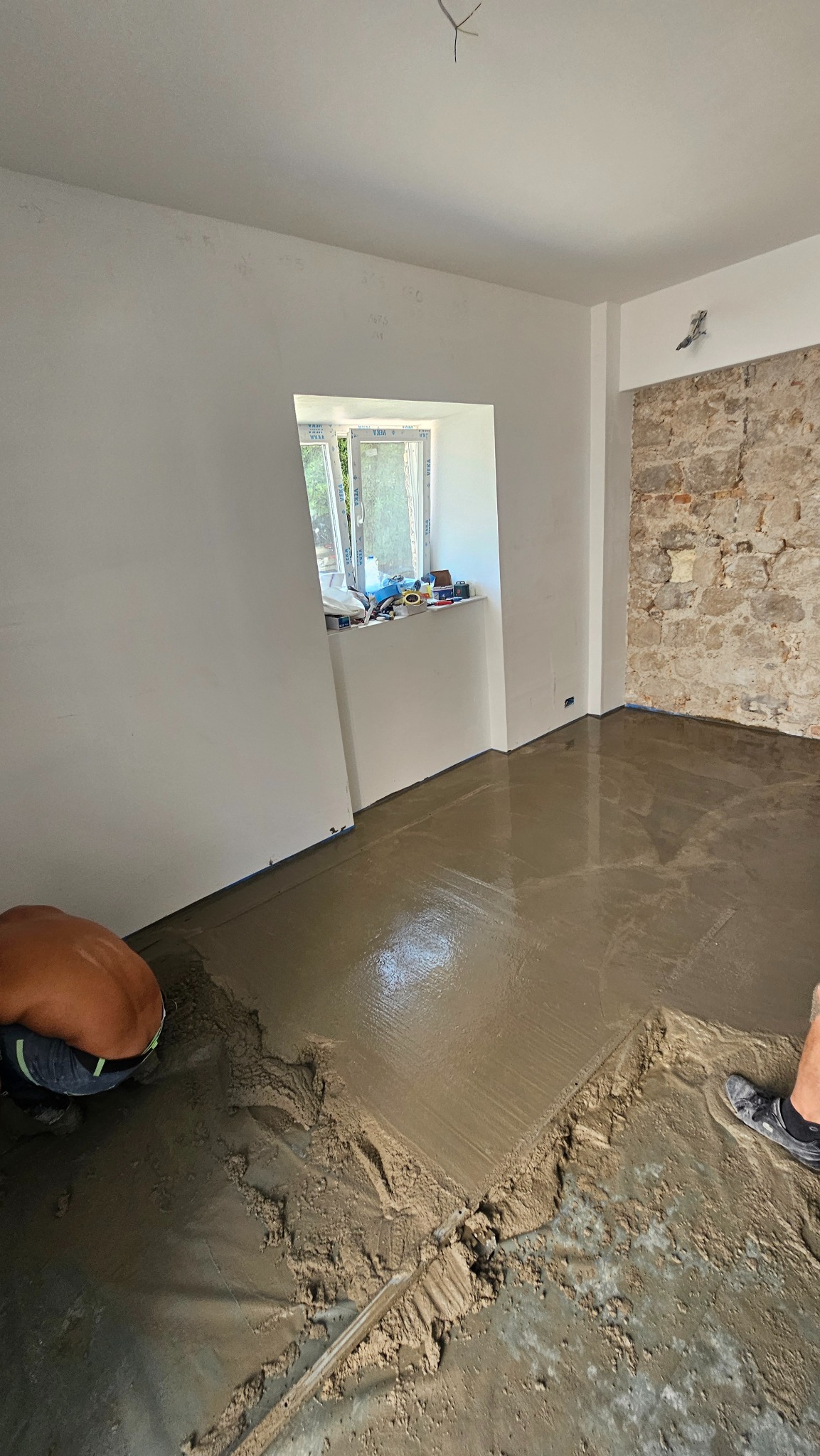 Wylewka podłogowa w jasnym pokoju z oknem i fragmentem ściany z kamienia. Pracownik w trakcie rozprowadzania masy. Widoczne narzędzia i folia ochronna.