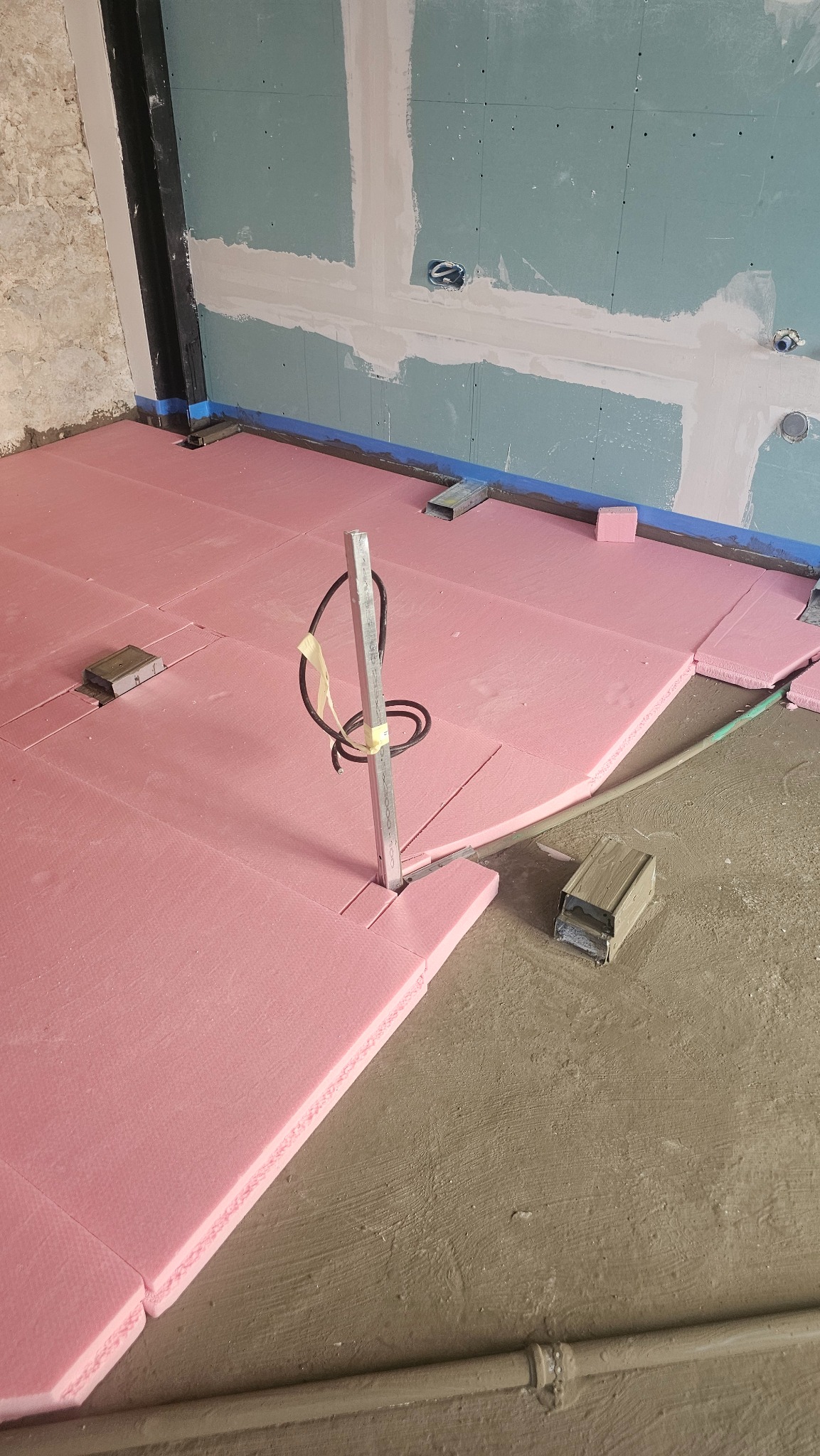 Różowa izolacja podłogowa EPS ułożona na szarym betonie, z metalowymi elementami mocującymi i pionową rurką z kablami. Ściany w surowym stanie, częściowo otynkowane.