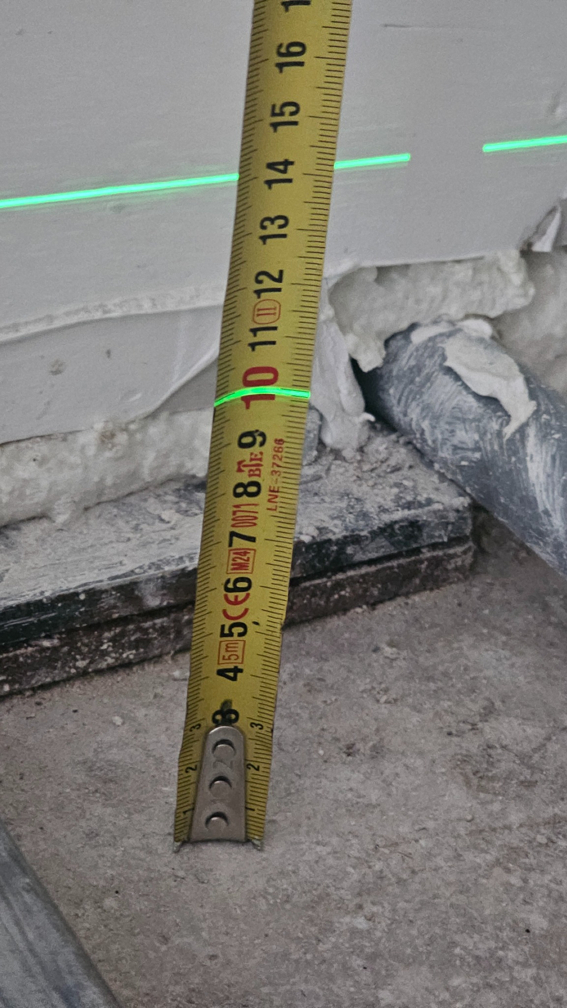 Pomiar poziomu laserowego z użyciem miarki zwijanej podczas prac remontowych. Widoczna instalacja i pianka montażowa.