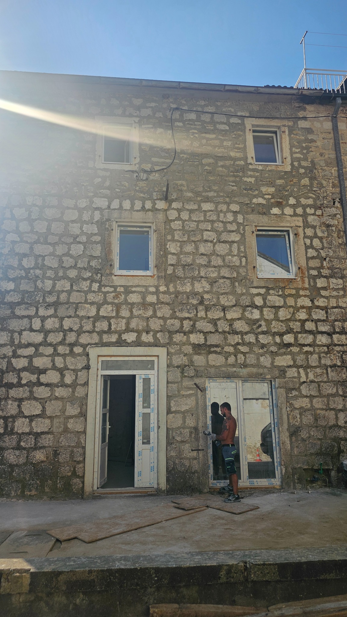 Kamienny budynek w trakcie renowacji: nowe okna i drzwi z PCV, pracownik w trakcie montażu. Słoneczny dzień, widoczne ślady prac budowlanych.