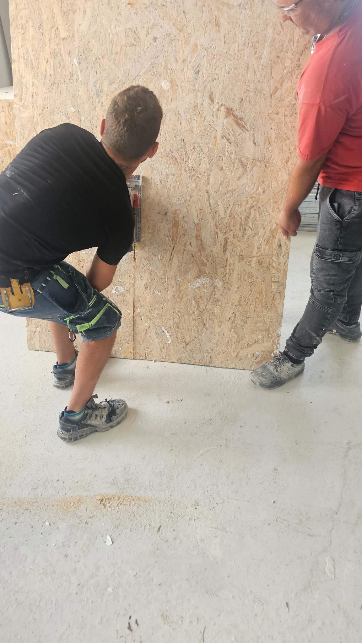 Dwóch monterów przy pracy z płytą OSB. Jeden mierzy, drugi przytrzymuje. Widoczne narzędzia i fragmenty podłogi z resztkami materiału.