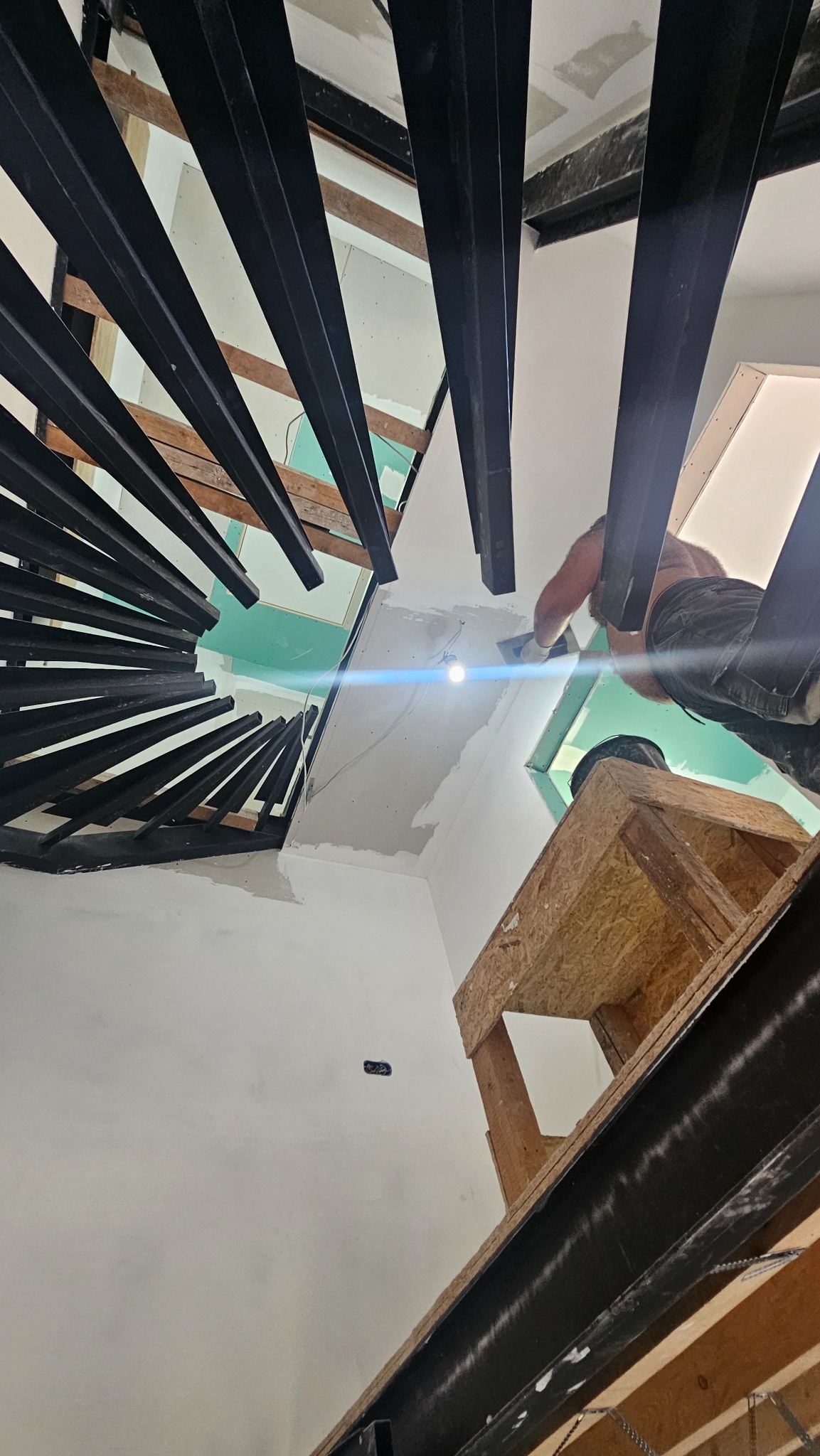 Remont klatki schodowej: widok z dołu na pracownika szpachlującego sufit przy schodach spiralnych z czarnymi elementami konstrukcyjnymi i drewnianym rusztowaniem.