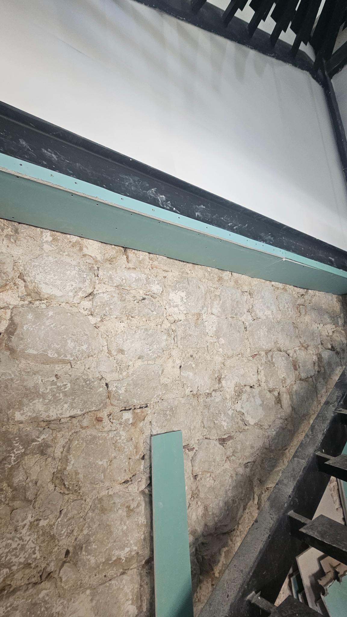 Fragment ściany z surowego kamienia, częściowo zakrytej płytą gipsowo-kartonową, z widoczną konstrukcją schodów w tle. Etap prac remontowych.