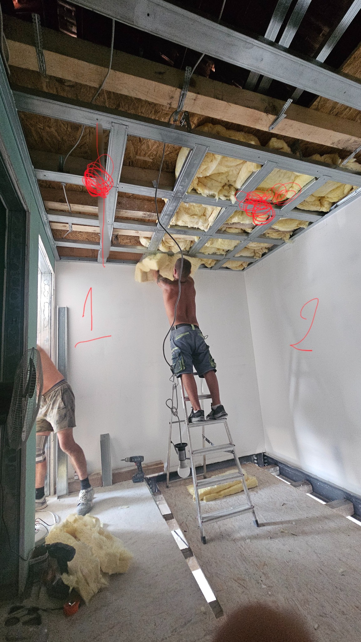Montaż izolacji stropu: mężczyzna na drabinie umieszcza wełnę mineralną w metalowej konstrukcji sufitu podwieszanego. Widoczne narzędzia i fragmenty izolacji na podłodze.