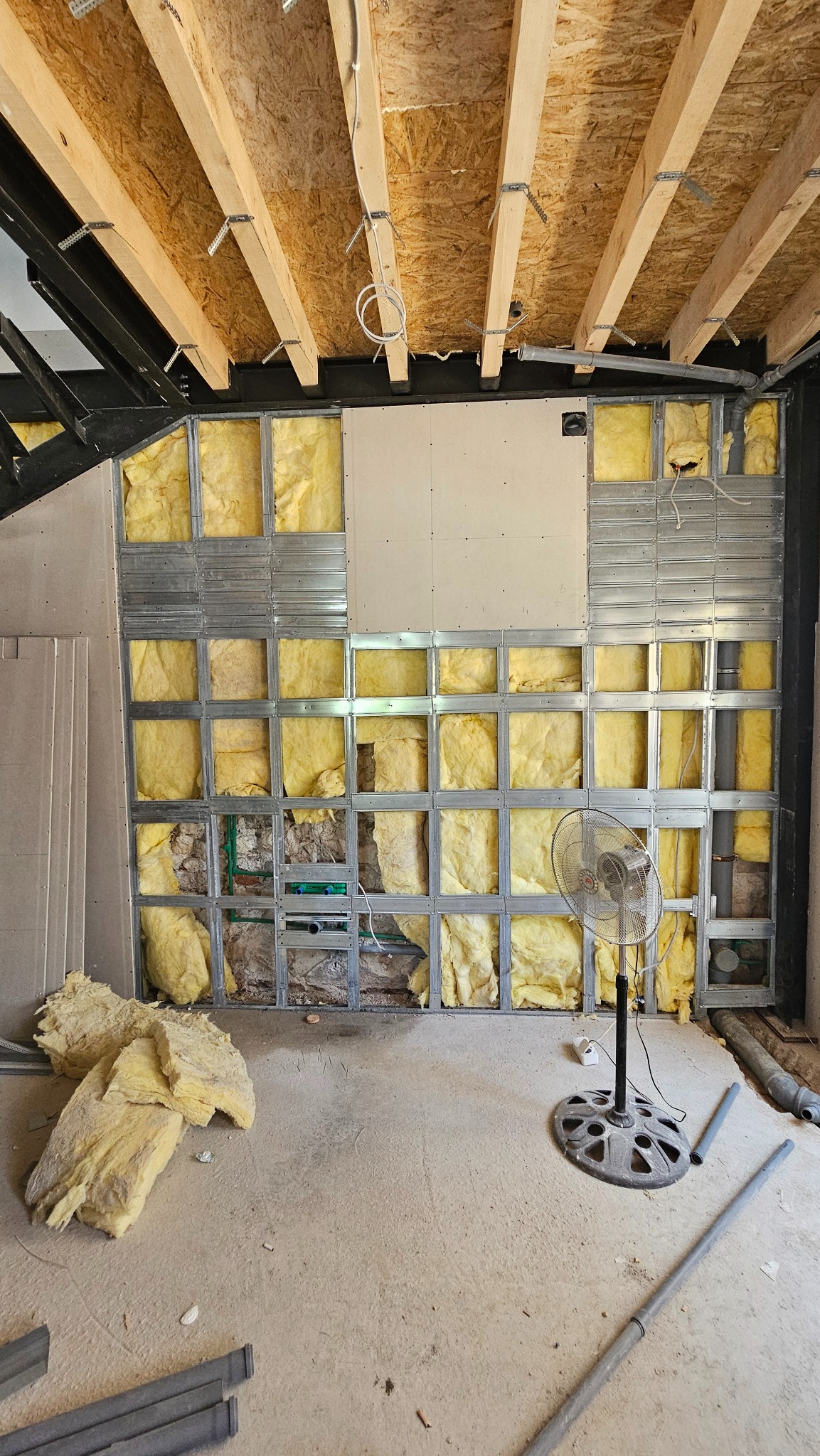 Wnętrze w trakcie remontu: metalowa konstrukcja ściany z izolacją z wełny mineralnej, widoczne rury instalacji, wentylator stojący na podłodze, surowy strop z drewnianych belek.