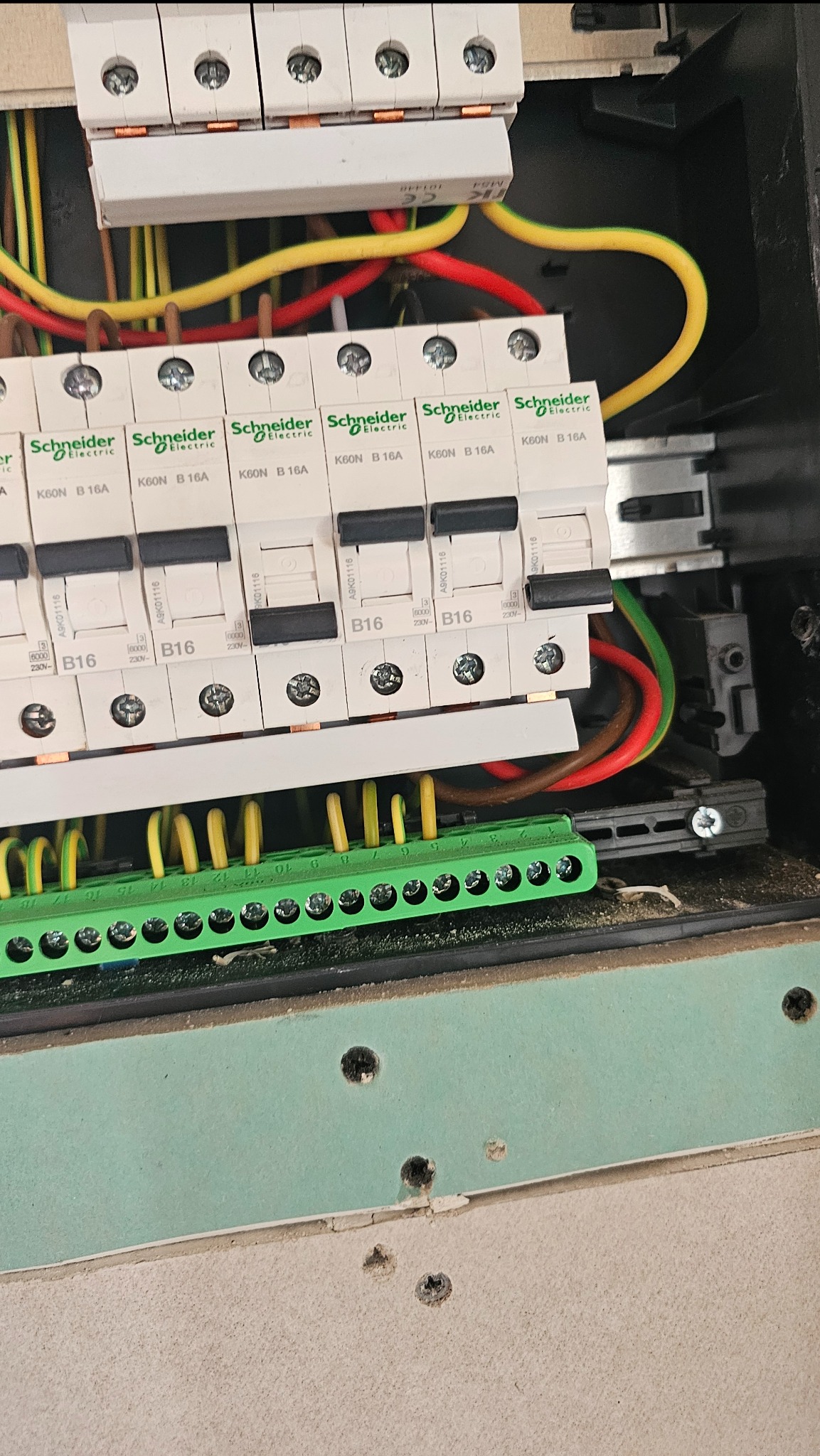 Rozdzielnica elektryczna z bezpiecznikami Schneider Electric K60N B16A i przewodami w kolorach żółtym, czerwonym i zielonym. Widoczne ślady montażu na ścianie.