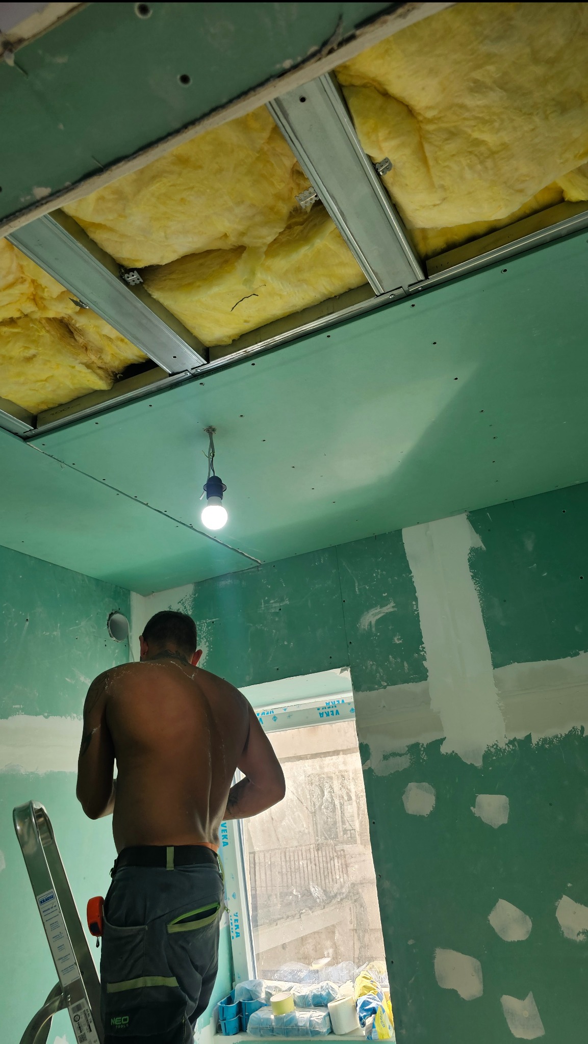 Montaż sufitu podwieszanego z płyt gipsowo-kartonowych i ociepleniem wełną mineralną. Widoczny pracownik podczas szpachlowania łączeń płyt.