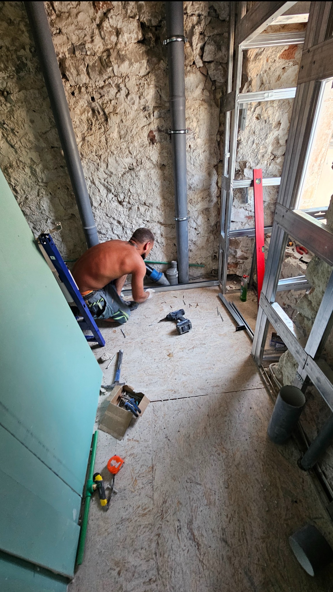 Hydraulik w trakcie pracy nad instalacją w pomieszczeniu z surowymi ścianami. Widoczne rury, narzędzia i konstrukcja szkieletowa ściany.