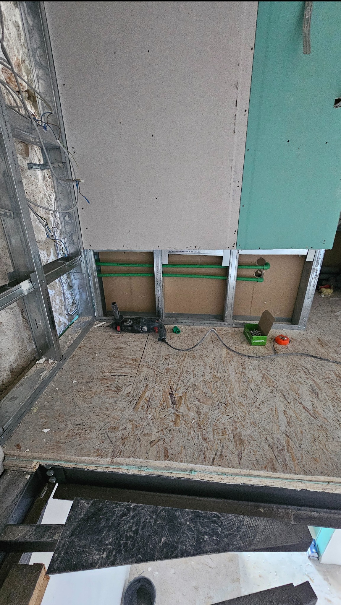 Wnętrze w trakcie remontu: widoczne płyty gipsowe, metalowa konstrukcja, instalacja hydrauliczna i narzędzia na podłodze z płyty OSB. Etap prac wykończeniowych.