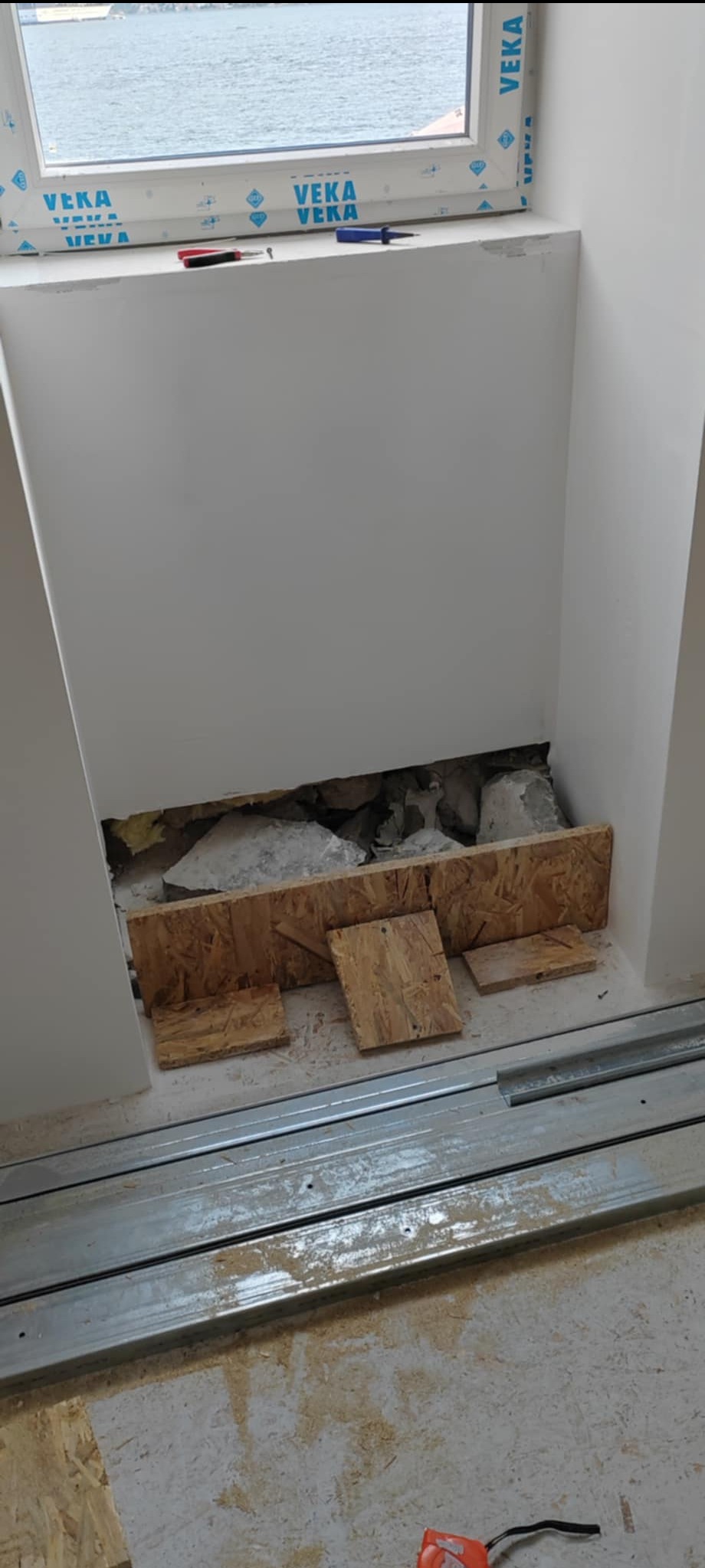 Wnęka okienna w trakcie remontu. Widoczne fragmenty starego muru, płyty OSB i narzędzia. Okno z widokiem na wodę. Prace przygotowawcze do wykończenia wnęki okiennej.