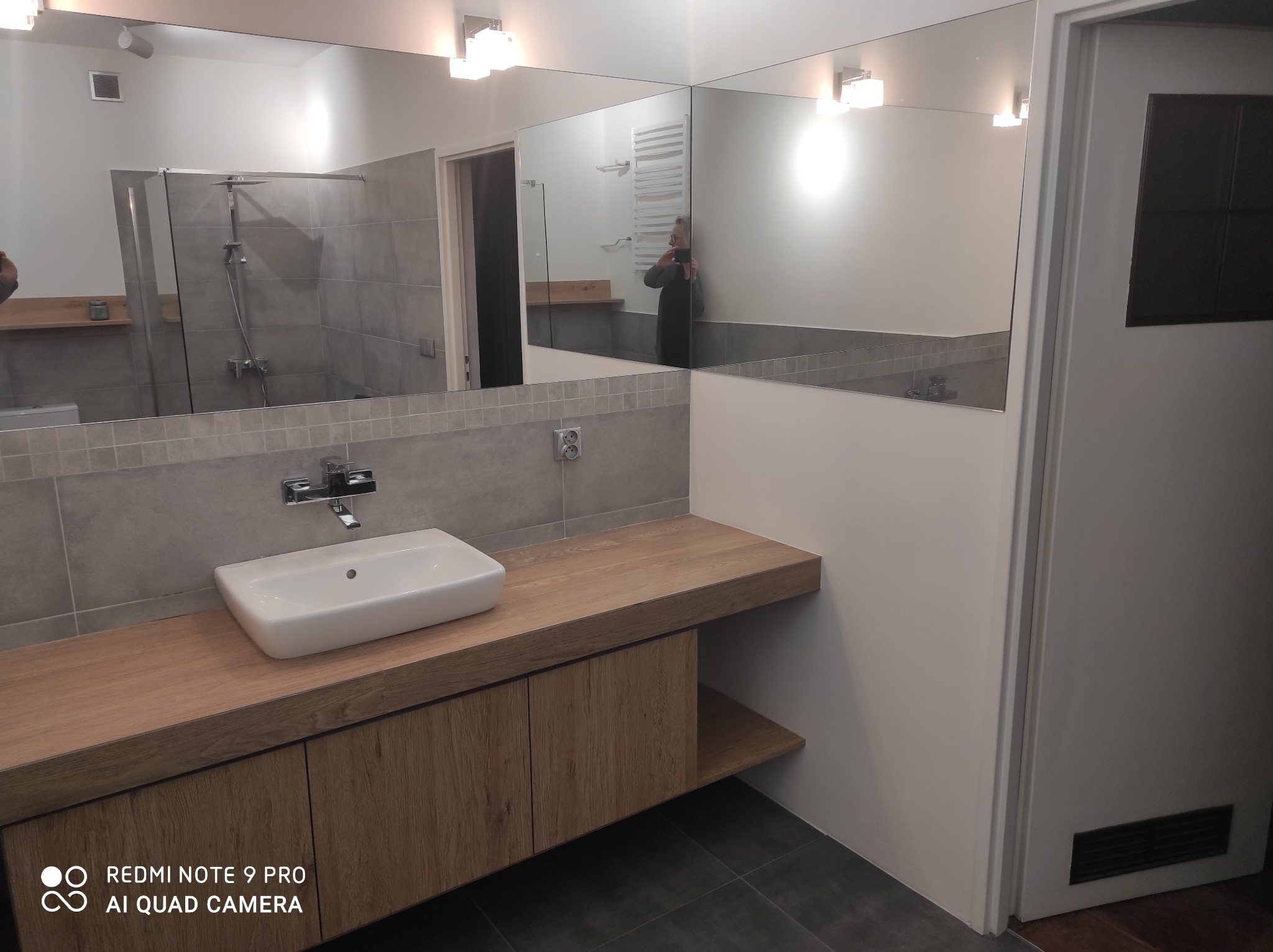 Nowoczesna łazienka po remoncie: biała umywalka na drewnianym blacie, szare płytki, duże lustro z oświetleniem. Widoczna kabina prysznicowa i grzejnik.