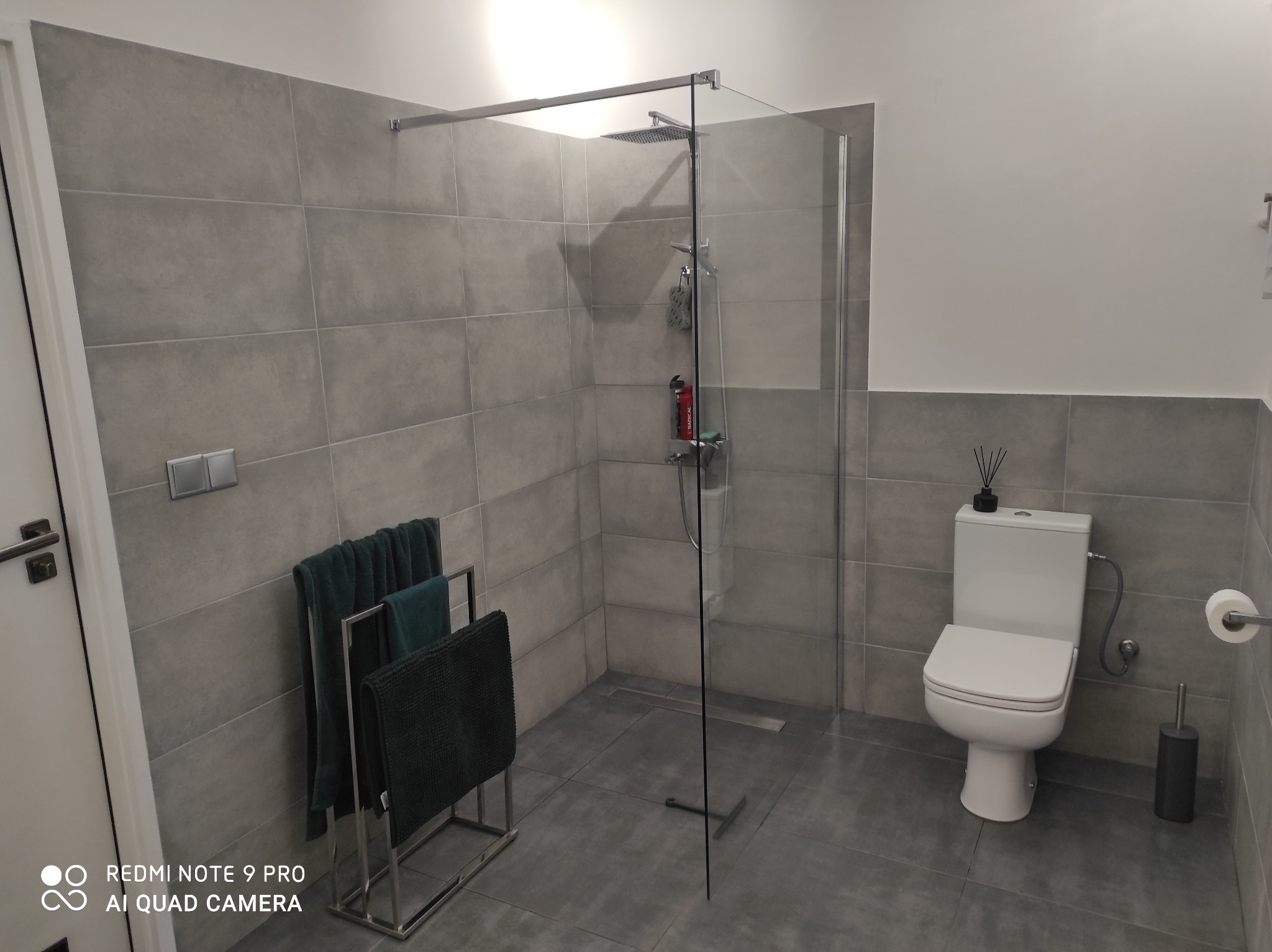 Nowoczesna łazienka z szarymi płytkami, kabiną prysznicową typu walk-in, toaletą i metalowym wieszakiem na ręczniki z zielonymi ręcznikami. Minimalistyczny design.