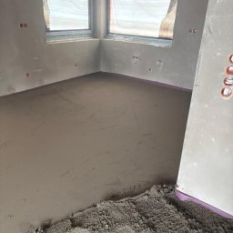 Posadzki Maszynowe - Posadzki Przemysłowe - ZAMBUD - Świeża, szara posadzka w surowym wnętrzu z oknem. Widoczne ślady stóp w materiale. Różowa izolacja przy ścianach. Gniazdka elektryczne na ścianach.