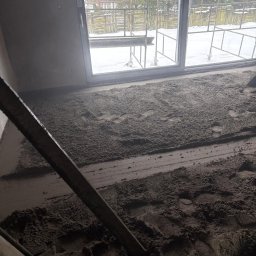 Posadzki Maszynowe - Posadzki Przemysłowe - ZAMBUD - Szara, świeżo wylana posadzka z widocznymi śladami stóp, częściowo wyrównana łatą. Za oknem rusztowanie i zimowy krajobraz.