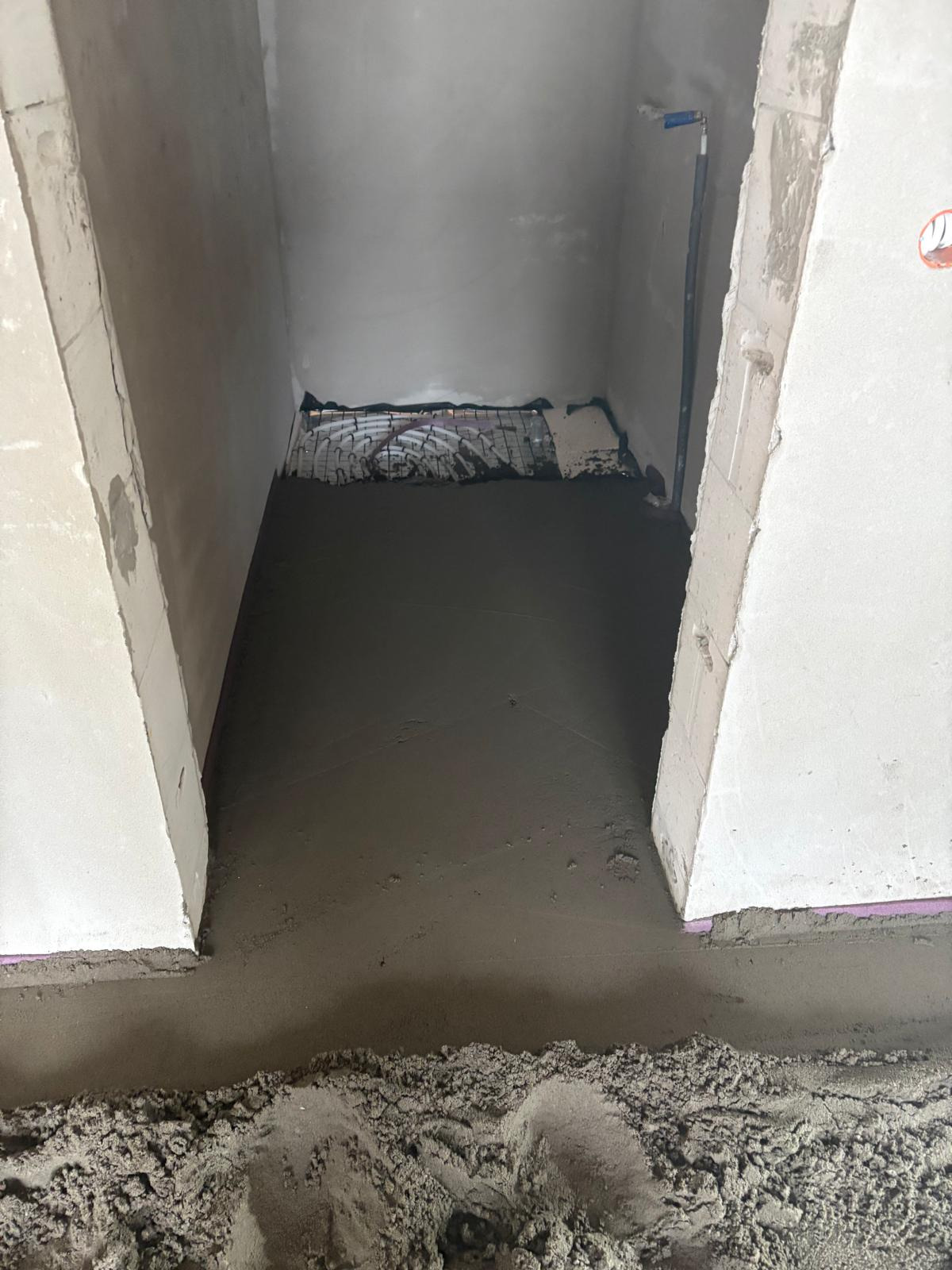 Świeżo wylana, gładka posadzka w pomieszczeniu z widoczną siatką zbrojeniową w tle. Surowe ściany i widoczne ślady prac budowlanych.