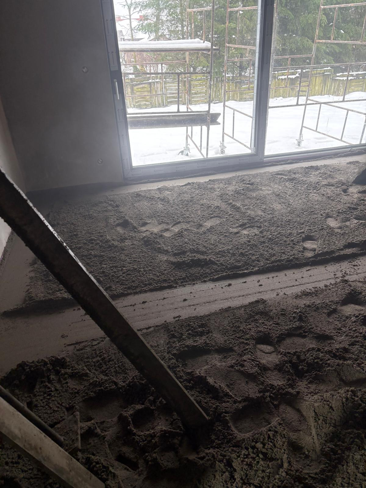Szara, świeżo wylana posadzka z widocznymi śladami stóp, częściowo wyrównana łatą. Za oknem rusztowanie i zimowy krajobraz.