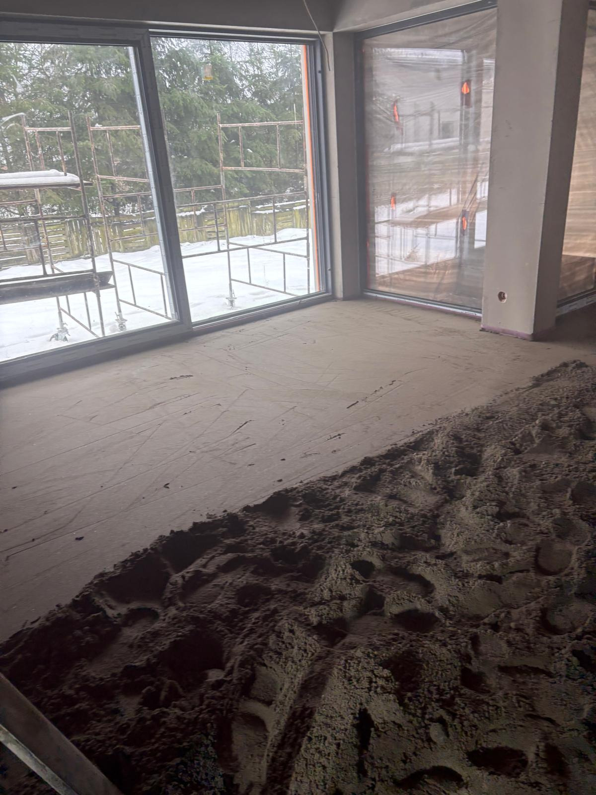 Wnętrze w trakcie prac posadzkarskich: częściowo rozprowadzona masa na podłożu, duże okna z widokiem na zewnątrz, rusztowania za oknem.