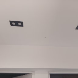 InstEl Jarosław Szulc - Sufit z dwoma czarnymi, kwadratowymi oprawami oświetleniowymi z dwoma okrągłymi otworami każda, rozmieszczonymi symetrycznie w białym suficie.