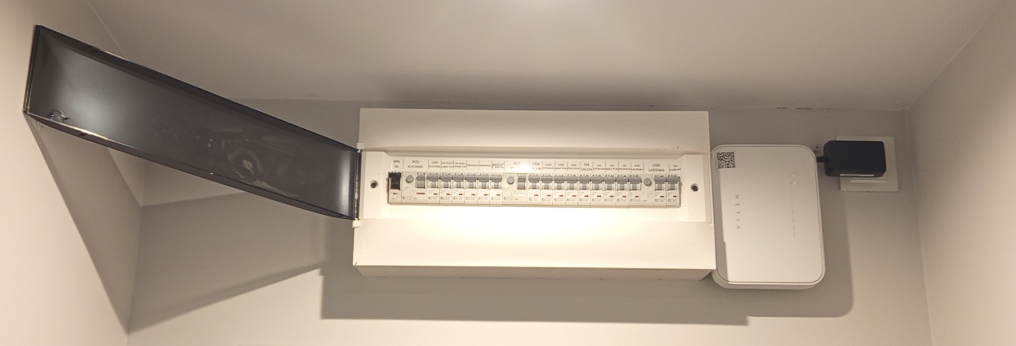 Rozdzielnica elektryczna z otwartą klapą, zamontowana na ścianie wewnątrz pomieszczenia. Obok widoczny router NETIA z zasilaczem. Białe ściany i sufit.