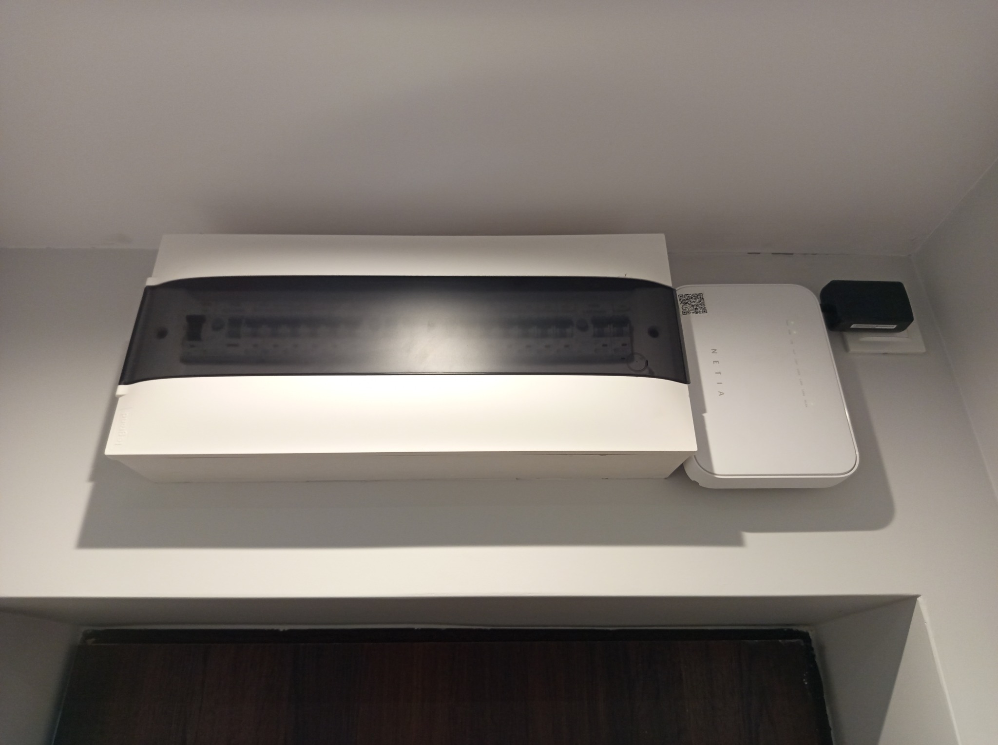 Biała skrzynka rozdzielcza z przezroczystym panelem na ścianie, obok router NETIA i gniazdko. Widok na górną część drzwi. Minimalistyczne wnętrze.