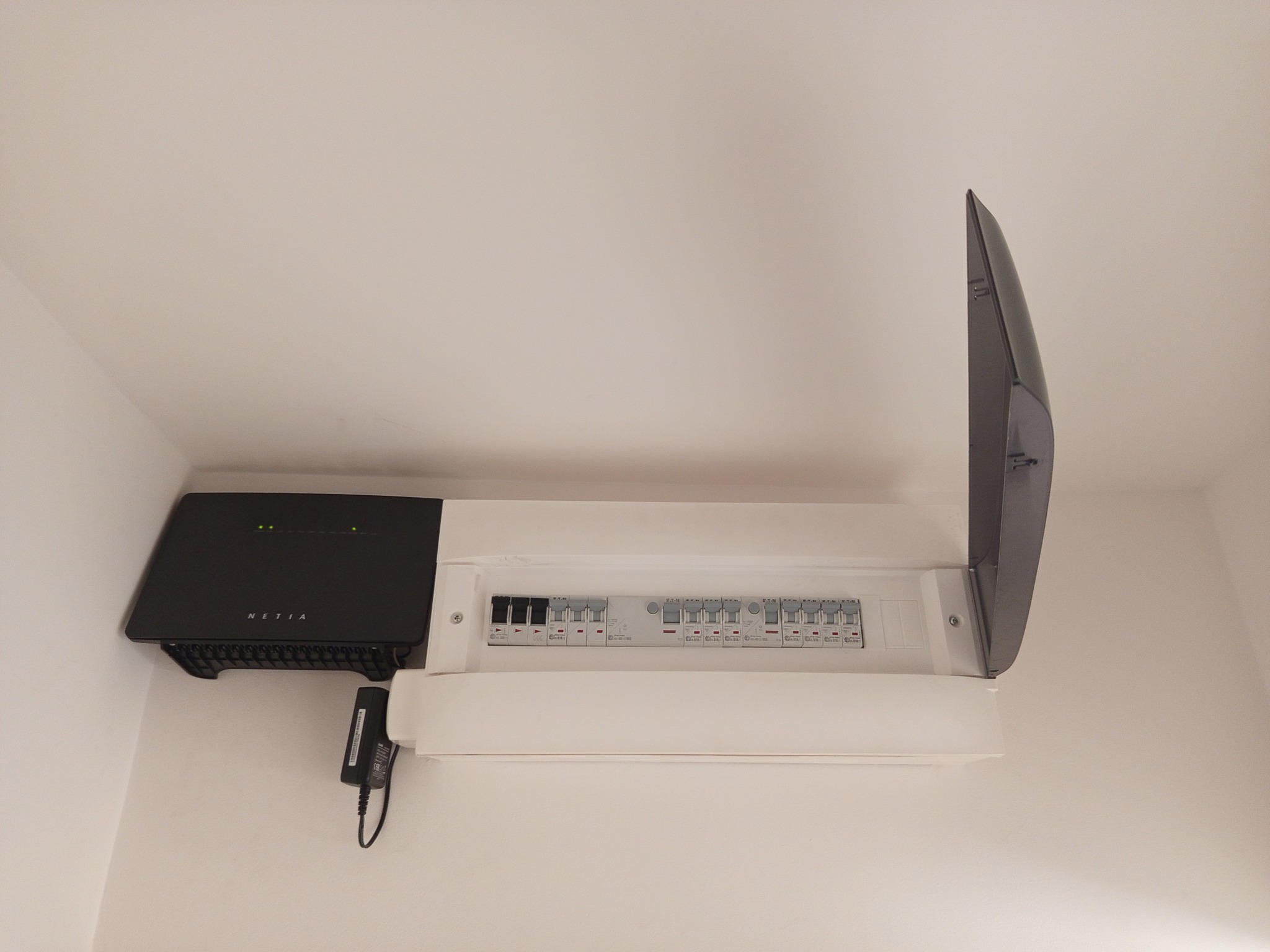 Biała skrzynka rozdzielcza z bezpiecznikami, zamontowana na ścianie, obok czarny router Netia z zasilaczem. Widok z dołu, pokrywa skrzynki otwarta.