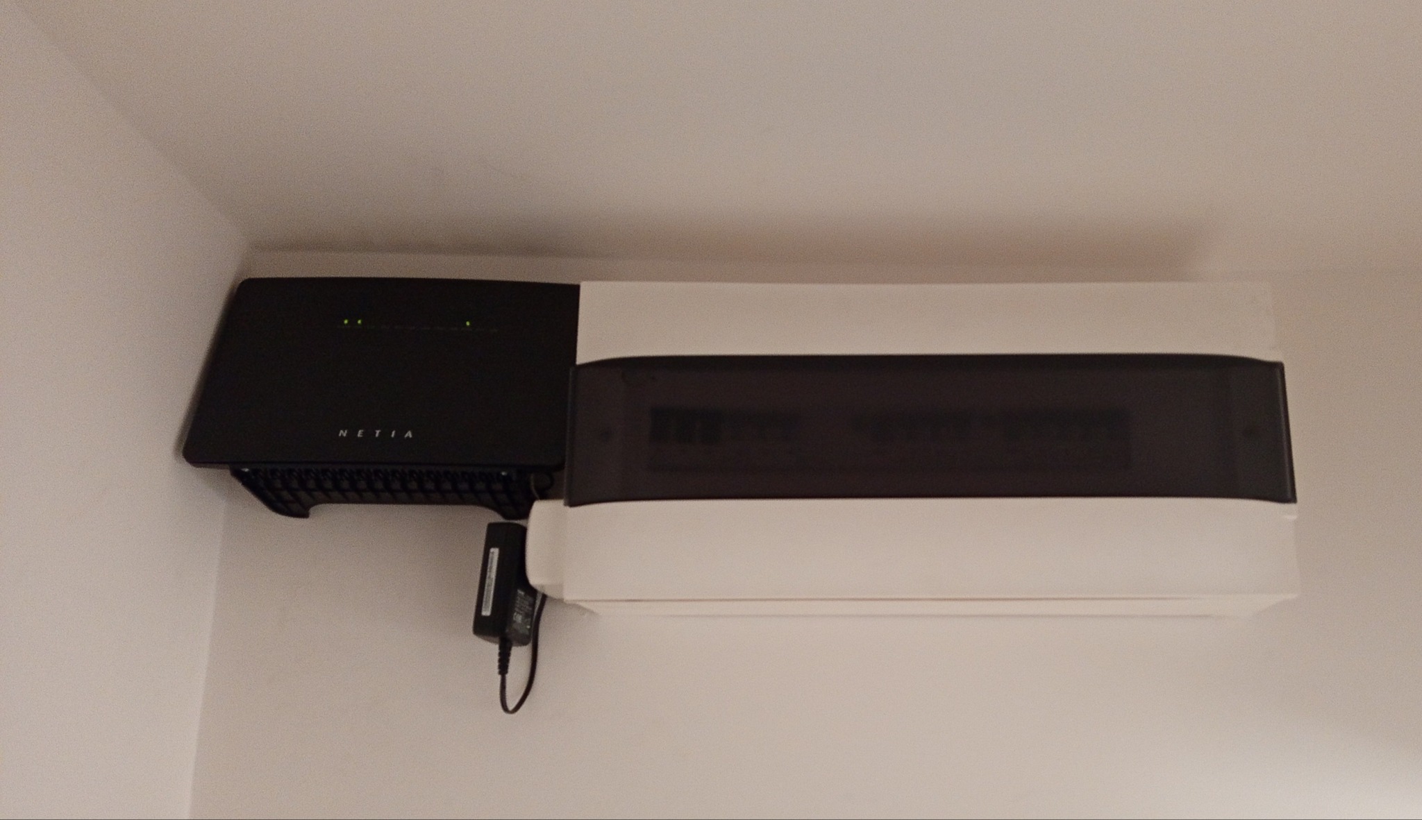 Biała skrzynka rozdzielnicza z czarnym panelem i routerem Netia zamontowana na ścianie. Widoczne diody sygnalizacyjne i zasilacz. Minimalistyczny design.