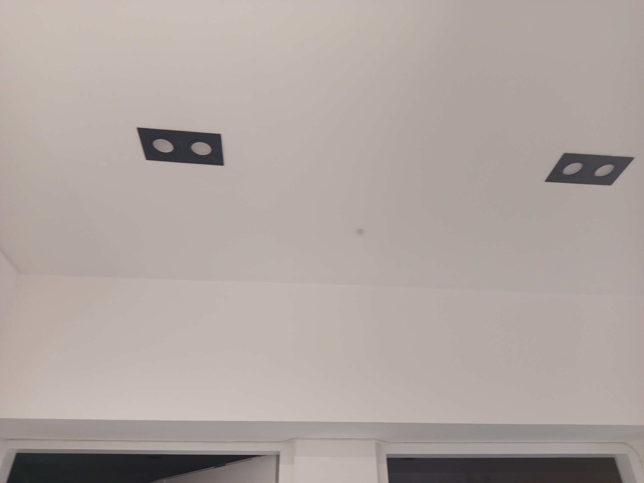 Sufit z dwoma czarnymi, kwadratowymi oprawami oświetleniowymi z dwoma okrągłymi otworami każda, rozmieszczonymi symetrycznie w białym suficie.