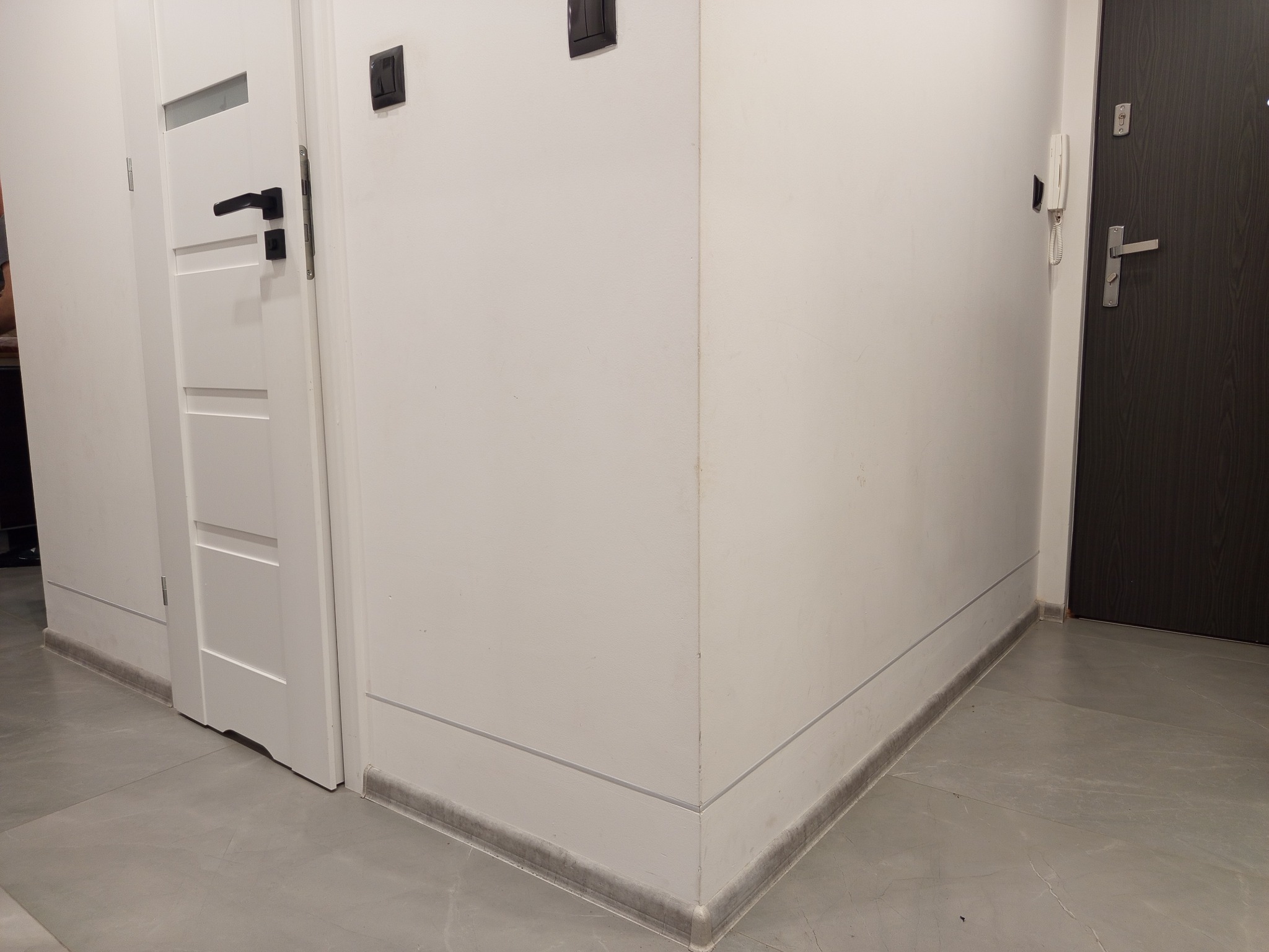 Wnętrze korytarza po remoncie: białe ściany z czarnymi włącznikami, szara podłoga, białe drzwi z czarną klamką i ciemne drzwi wejściowe z domofonem.