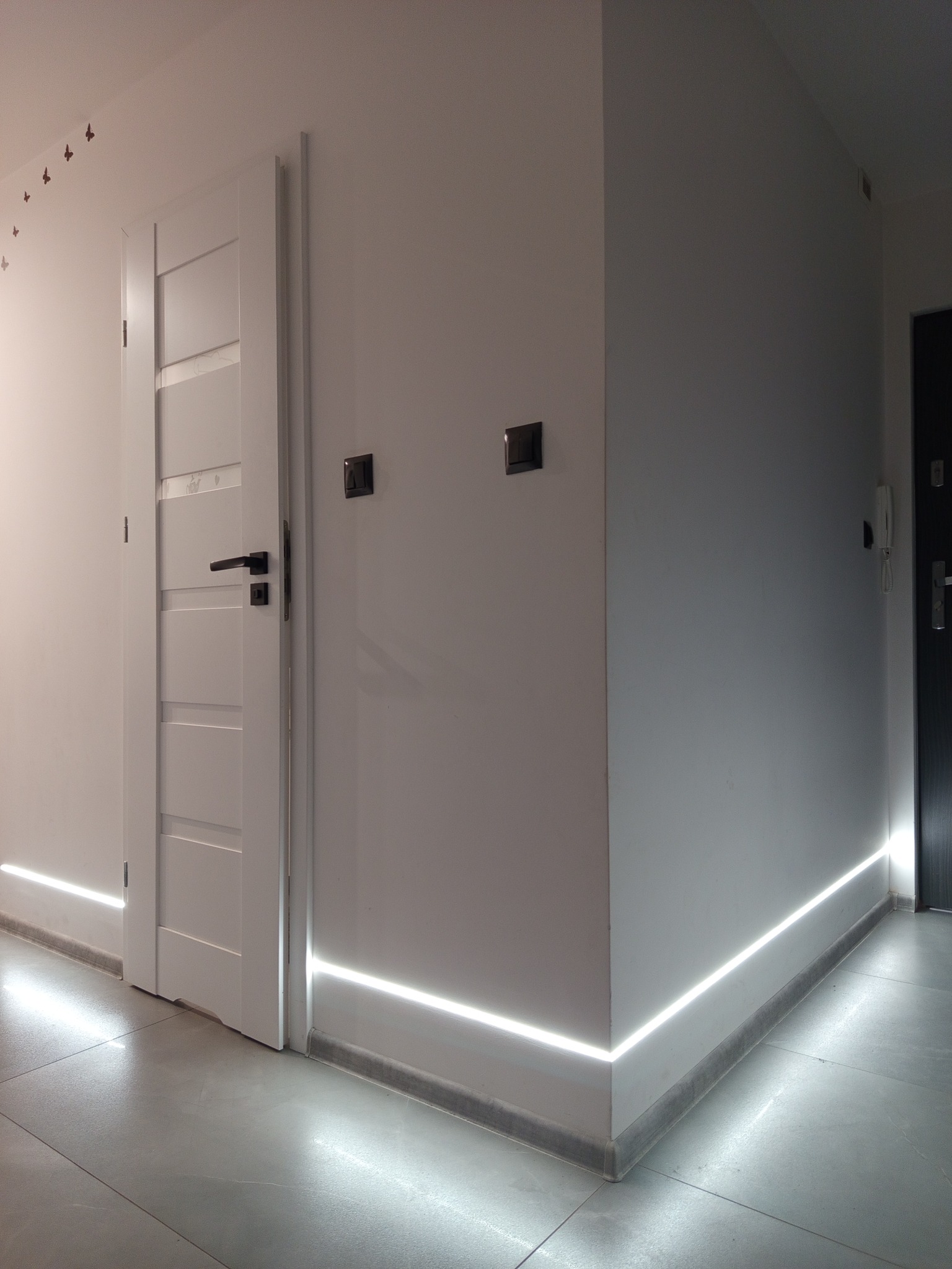 Nowoczesne oświetlenie LED przypodłogowe w korytarzu, subtelnie rozświetlające przestrzeń. Widoczne białe ściany, szara podłoga i drzwi. Minimalistyczny design.