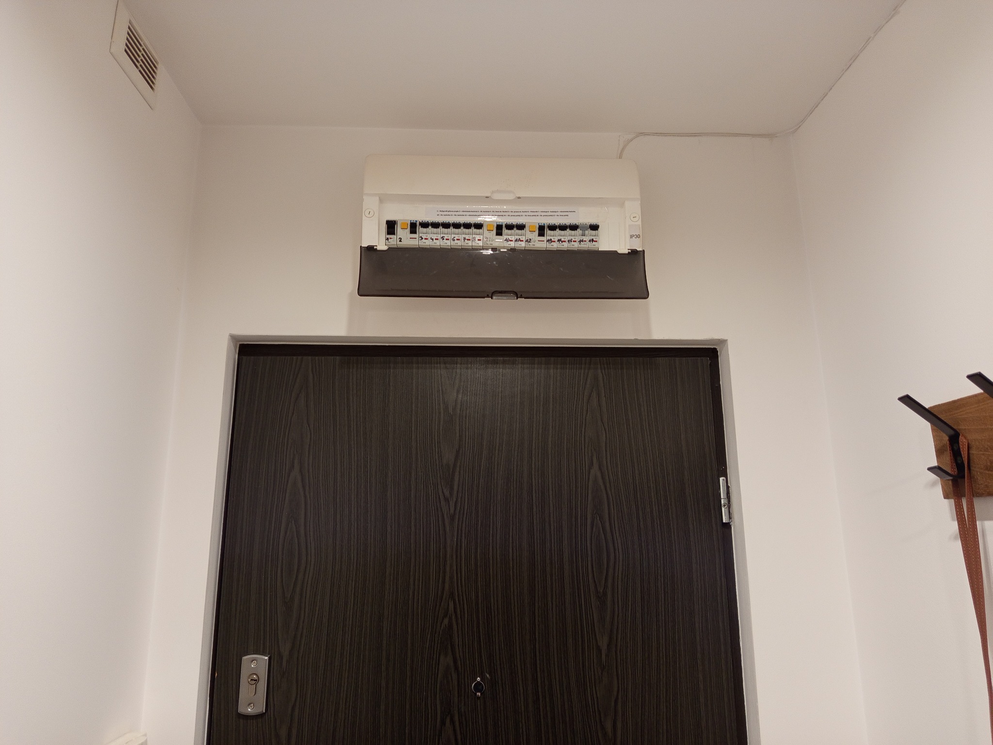 Otwarta skrzynka bezpiecznikowa z widocznymi bezpiecznikami, zamontowana nad ciemnymi drzwiami w białym korytarzu. Widoczny fragment wieszaka na ścianie.