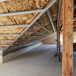 Marbud - Poddasze z izolacją z wełny mineralnej i metalową konstrukcją pod płyty gipsowo-kartonowe. Widoczne drewniane elementy konstrukcyjne i surowy beton na podłodze.