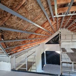 Marbud - Skośny sufit poddasza w trakcie prac wykończeniowych: widoczna izolacja, metalowa konstrukcja pod płyty gipsowe i rusztowania. Prace na wysokości.