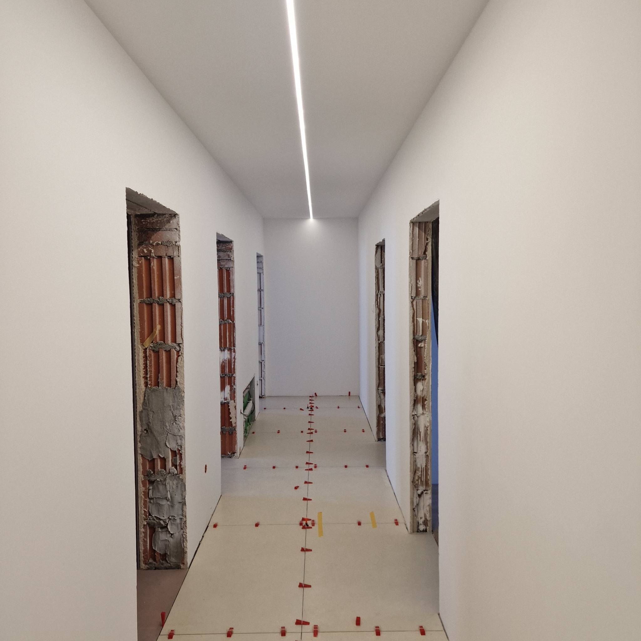 Korytarz w trakcie remontu: świeżo pomalowane ściany, surowa cegła w ościeżnicach i ułożone płytki podłogowe z elementami poziomowania. Oświetlenie liniowe LED.